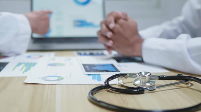 Doctors consulting together and planning treatment in a modern medical office, reviewing healthcare data, charts, and reports on a laptop for clinical decision making and patient care.