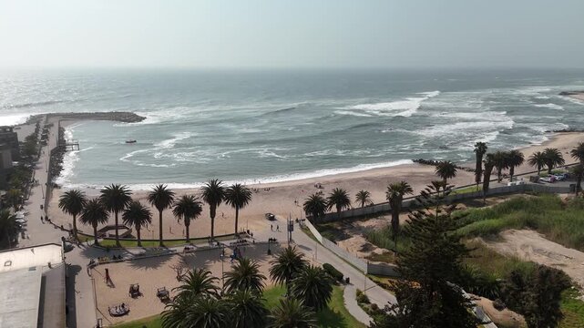 Cinematic aerial footage of the rough Atlantic Ocean waves washing onto the sandy beach beside a beautiful palm-lined promenade in Swakopmund, Namibia.