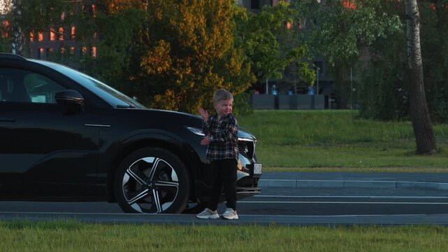 Child in plaid shirt interacts with black electric car, showcasing playful gestures in a grassy area during sunset with urban background