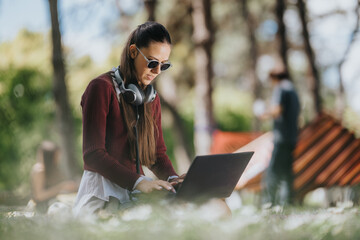 Fototapeta premium Young woman sitting on grass working on a laptop while wearing headphones and sunglasses. A relaxed outdoor study scene shows her focused on typing in a park setting.