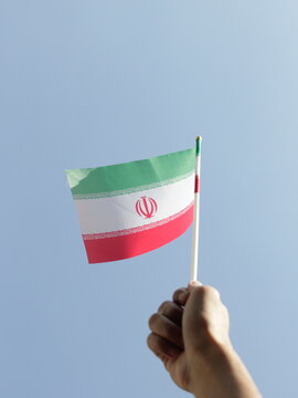 A close-up of a hand waving a miniature Iran flag with a bright blue sky in the background.