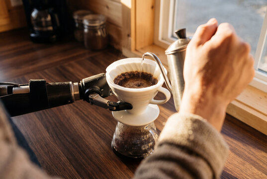 Person with prosthetic arm pouring hot water over coffee grounds in dripper on wooden kitchen counter.