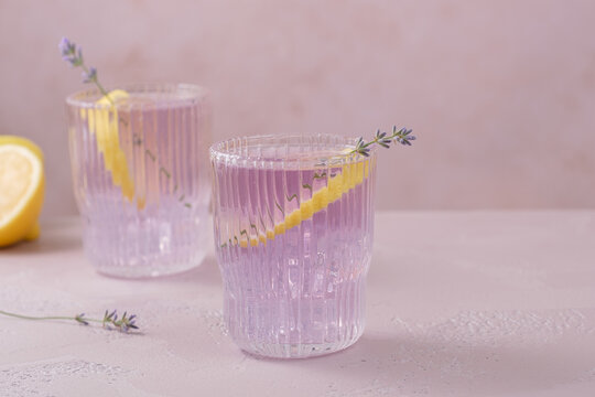 Two glasses of lavender lemonade are placed on a light table. Each glass has a lemon slice and a lavender sprig on top. A lemon halves is nearby.