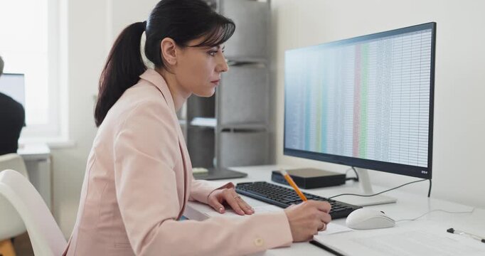 Businesswoman in office typing document and paperwork. Focused manager reviews spreadsheet data, taking notes daily at a professional workplace. Office productivity and computer planning concept.
