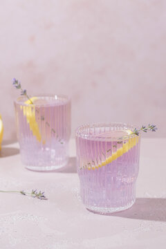 Two glasses of lavender lemonade are placed on a light table. Each glass has a lemon slice and a lavender sprig on top. A lemon halves is nearby.