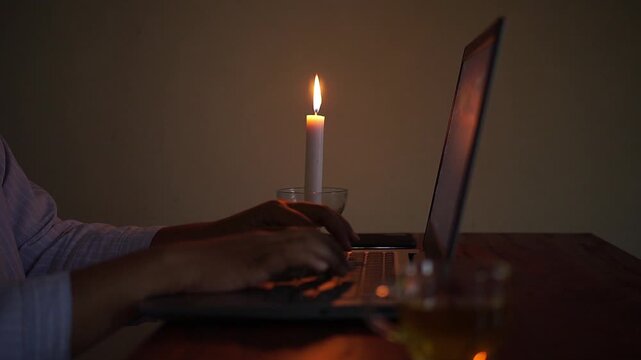 Hand typing on a laptop beside a lit candle during a power outage. Cozy home office scene representing remote work, productivity, business continuity, and working in low light