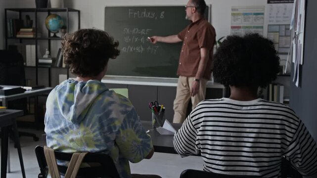 Middle aged Caucasian male teacher explaining algebra equations on blackboard to diverse teenagers in classroom. Clip for promoting high school tutoring services or e-learning campaigns