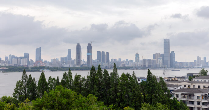 A wide Wuhan city skyline across a broad river framed by leafy trees in the foreground, featuring modern high-rise buildings, boats on the water and a moody overcast sky for an urban waterfront scene