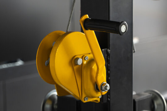Close-up of a yellow hand winch pulley with a black crank handle mounted on a metal frame, illustrating industrial lifting equipment, workshop hardware, rigging, and mechanical detail for maintenance