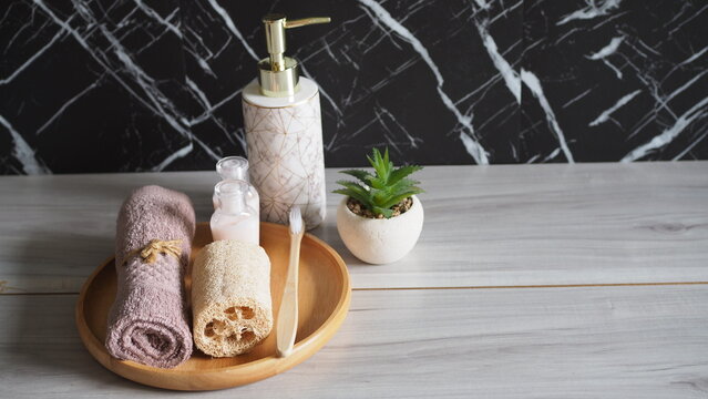 Luxury eco-friendly bathroom setup with a bamboo toothbrush, natural loofah, and rolled pink towel on a wooden tray, paired with a white marble ceramic soap dispenser. Side view on a light gray wood 