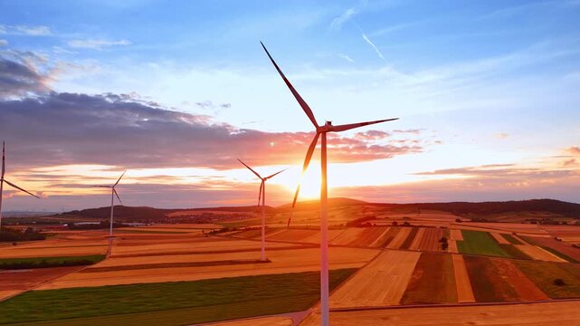 Wind farm turbines silhouetted against orange rural sunset. Aerial view shows four towers over geometric fields, with sunlight passing through the nearest rotor.