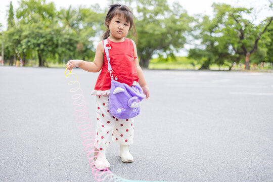 Adorable Asian Girl Playing with a Rainbow Slinky Spring Toy Outdoors,A cute young Asian girl standing in a park, holding and playing with a vibrant, colorful rainbow-colored slinky spring toy.