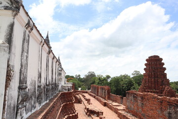 Fototapeta premium Ancient Temple Ruins under Sky: The weathered white wall of an ancient temple runs alongside decaying brick ruins, set against a backdrop of lush green trees and a partly cloudy sky.