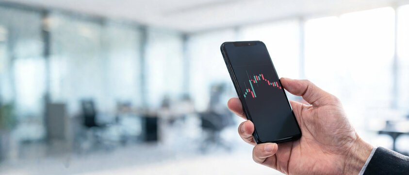 Primer plano de la mano de un empresario sosteniendo un smartphone con un gr&aacute;fico financiero de trading de velas en una oficina borrosa. Formato panor&aacute;mico ideal para banner.