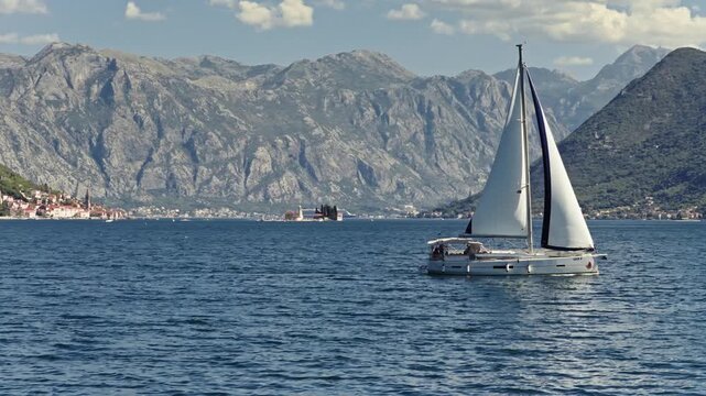 aerial view of a white sailboat under full sail crossing the deep blue water of the bay of kotor, passing the island church of our lady of the rocks and perast below steep mountains, montenegro