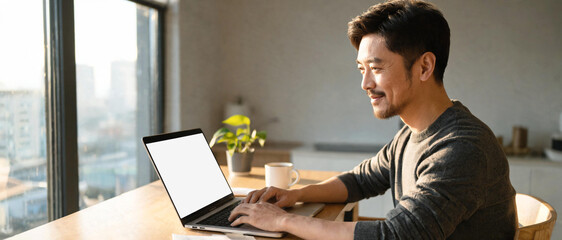Profesional asiático sonriente trabajando con su laptop con pantalla blanca para mockup en un escritorio de madera junto a la ventana. Concepto de teletrabajo y negocios. © Othoniel