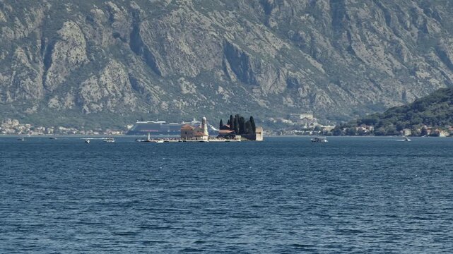 aerial view of the island church of our lady of the rocks and the town of perast in the bay of kotor, with small boats on the water and steep mountains behind, montenegro