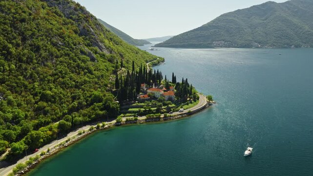 aerial view of a stone waterfront estate with orange tile roofs and tall cypress trees on a forested headland in the bay of kotor, a coastal road and a mussel farm on calm water, montenegro