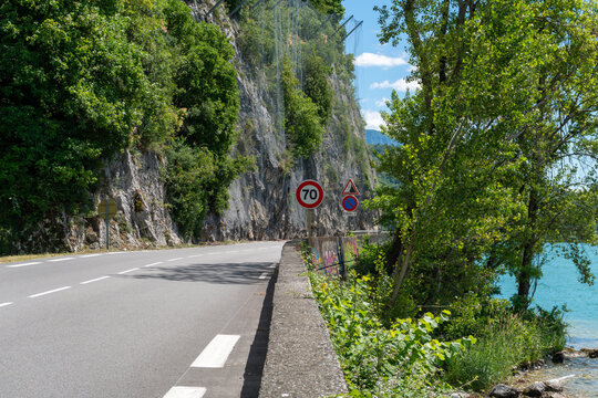 Curving road beside Lake Bourget in Savoie, France, running between turquoise water and a steep limestone cliff protected by rockfall netting. Traffic signs, retaining wall and dense vegetation illust