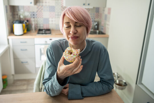 Pleased positive mature woman enjoying eating sweet glazed doughnut snack in kitchen, smiling female tasting high-calorie pastry dessert, junk food and sugar treats addiction, sweets indulgence.