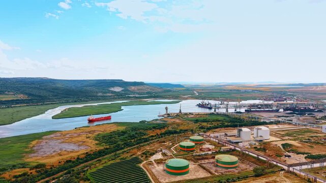 Flyover the tanks on the territory of industrial plant at daytime. Approaching the waterfront with cargo ship heading to the port.