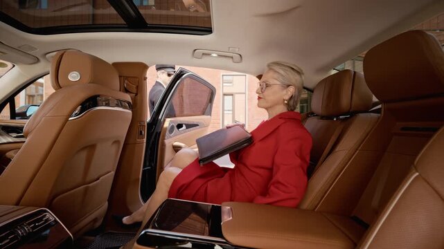 Mature Caucasian businesswoman getting into luxury car back seat opened by chauffeur, opening leather folder to review documents. Perfect for executive travel and corporate leadership videos