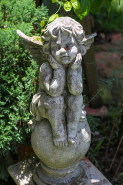 Weathered stone cherub statue sits thoughtfully in a lush garden setting
