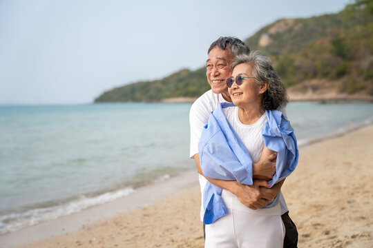 Happy Senior Asian Couple Embracing Loyally on Sandy Beach, Retirement Travel Concept