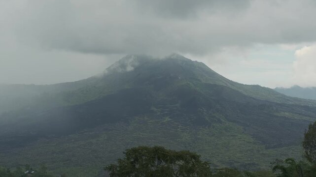 Time-lapse of Mount Batur volcano in Kintamani, Bali, with clouds and mist covering the peak, surrounded by dense green forest during daytime.