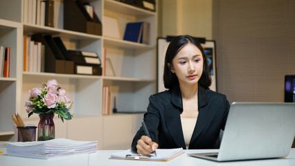 Fototapeta premium asian businesswoman working at her desk, focused on taking important notes on a notebook while using her laptop, ideal for business and corporate concepts.