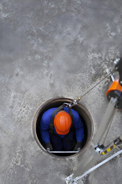 Overhead view of industrial worker in blue coverall and orange hard hat descending into round manhole confined space with safety tripod rescue system, confined space entry procedure concept