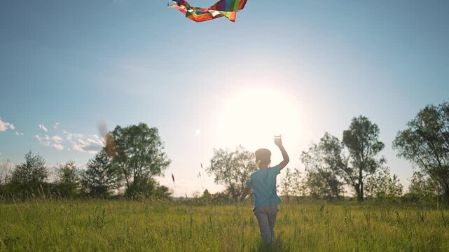 Rainbow kite flies in evening sky. Boy stands on meadow watching kite. Grass rustles under breeze. Sunlight wraps boy in nature calm. Kite colors glow rainbow. Meadow full of grass. Kite floats in sky