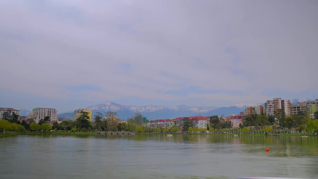 Timelapse: different modern apartment buildings and park on the shore of lake in Batumi - cloudy sky, overcast. People are kayaking on the lake. Moving clouds over the mountains - time lapse