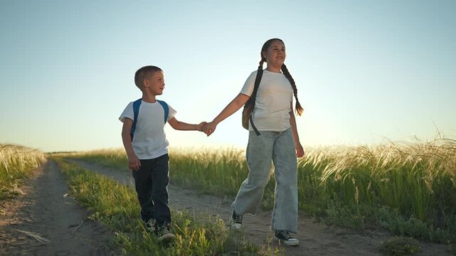 Brother sister holding hand walking across field. Child backpack steps beside sister nature path. Sunset warms grass around them. Brother sister enjoy calm field walk together under soft summer sky.