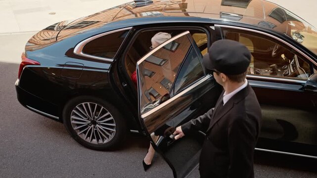 Caucasian male chauffeur opening door of luxury black sedan for senior Caucasian businesswoman talking on smartphone. Ideal for corporate travel and VIP service promotions