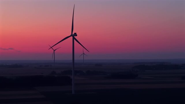 Wind turbines at a wind farm during dawn, featuring flashing red obstruction lights on the nacelles or towers and continuously rotating blades that convert wind into clean, renewable electrical energy