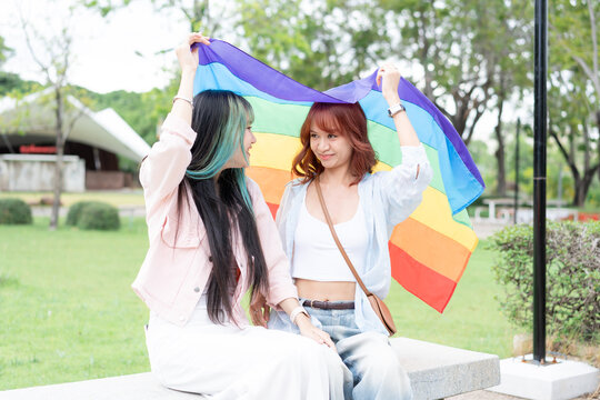 Beautiful young Asian lesbian couple holding rainbow pride flag over heads in public park. Romantic LGBT women looking at each other with love, freedom and equality concept.