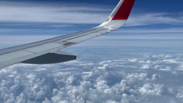 White airplane wing with red and white vertical stabilizer diagonally framed against dense white cumulus clouds and blue sky, captured from a high-angle view above the cloud layer during daytime.