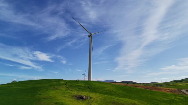 Drone footage captures multiple white wind turbines on rolling green hills under a blue sky with clouds in rural Spain, aerial perspective.