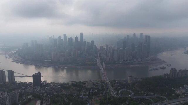 Cinematic drone shot slowly moving forward toward the densely developed Chongqing peninsula under heavy low clouds. The Yangtze River, Dongshuimen Bridge, high-rise skyline, river traffic and multilay