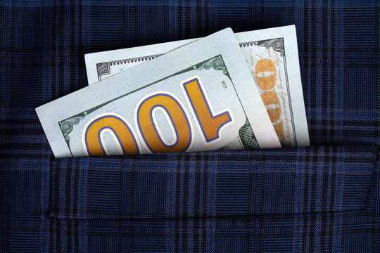 Close-up of two US hundred dollar bills sticking out of a blue plaid fabric pocket, symbolizing wealth, savings, or finance concepts.