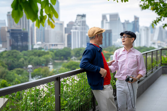 Happy Senior Asian Men Chatting on Balcony with City Park and Skyline Background