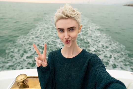 Young woman taking a selfie on a boat showing a peace sign with ocean wake background for social media banner and lifestyle design