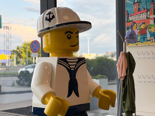 Fototapeta premium A giant lego-style sailor figure stands inside a shop by a bright window, creating a playful retail display with a nautical theme.
