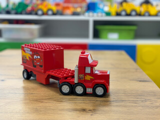 Fototapeta premium Red toy truck with a cartoon face on a wooden table, with a blurred classroom play area and colorful toys in the background