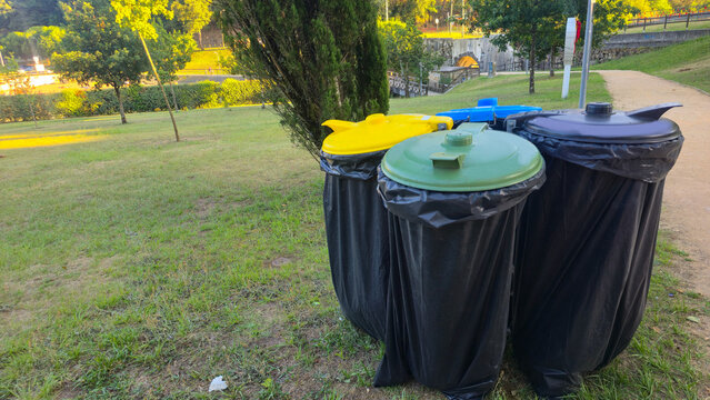 On a sunny day, trash bins for sorting waste line a pedestrian path in a green city park. Concepts: recycling, waste management, sustainability, cleanliness, public services, urban life.