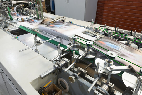 View of a folder gluer machinery system in operation at a printing plant for the creation of paper bags including various rollers and belts for a fast and automated assembly process in the factory.
