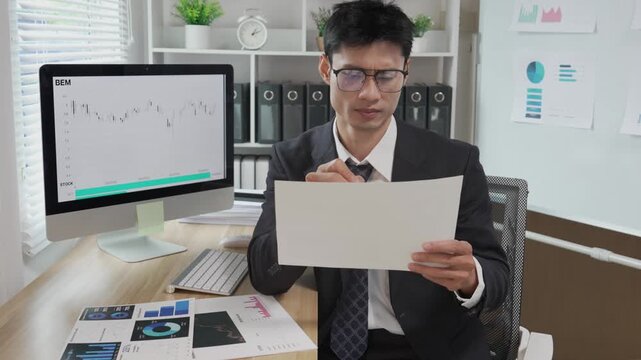 A stressed Asian businessperson holds documents and looks anxious about errors in a financial report amidst a stock market slump or financial losses.
