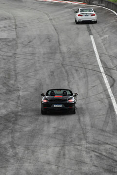 Black Porsche 718 Boxster and white BMW M4 (F82) driving on a tire-marked racetrack