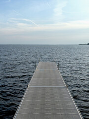 Fototapeta premium Long floating dock stretching into calm open water beneath a soft blue sky with wispy clouds.
